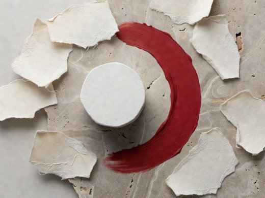 Overhead 3D render of an architectural flat lay with torn cotton paper, geometric plaster cylinder, and a curved sweep of crimson pigment on travertine
